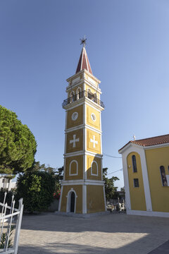 View of the yellow church bell tower with a red roof and white crosses under a clear blue sky in Argassi, Greece.