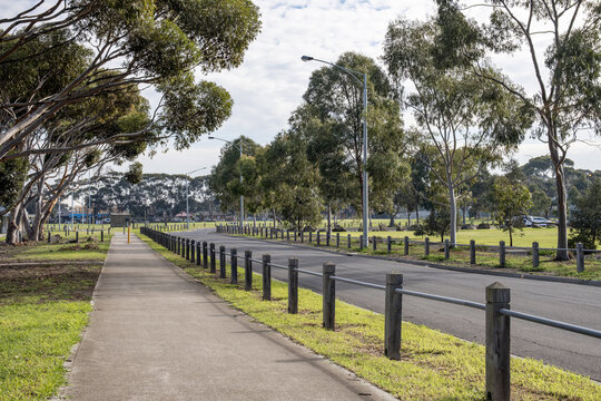 a concrete footpath running parallel to an access road with post and rail timber barriers, street lamps, eucalyptus trees, and grassy open parkland in suburban Melbourne, Australia.