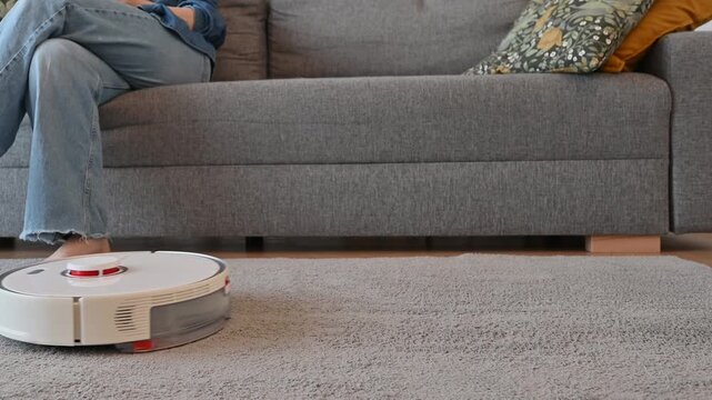 Smart vacuum cleaner cleans the carpet while woman rests on the sofa. A smart home, modern technologies,housekeeping concept