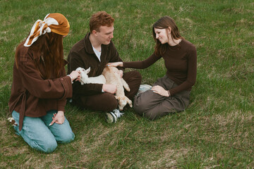 Obraz premium Casual outdoor scene with a family and friends petting a curious goat kid on a sunny grassy field, relaxed smiles, warm tones, gentle breeze, tender moment of countryside lifestyle