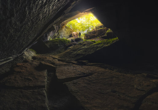 View of Cave di Pietra Molera with a person standing at the entrance illuminated by sunlight filtering through green foliage in the dark rocky cavern in Malnate, Lombardy, Italy.