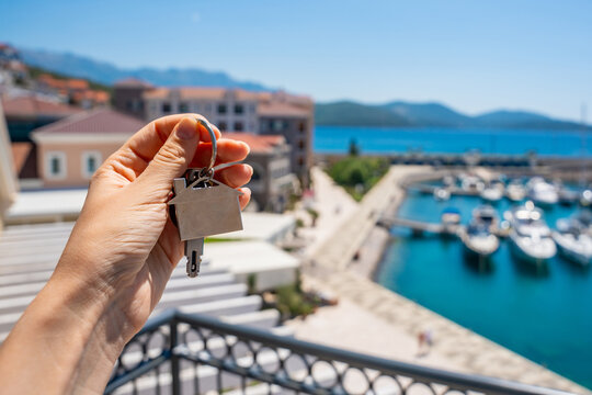 Hand holding a house key with a blurred seaside villa and marina in the background. Concept of buying coastal real estate, property investment and owning a vacation home.