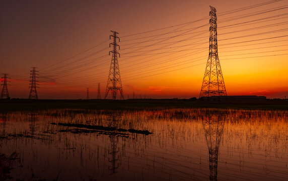 High voltage electricity transmission towers reflected in flooded field during vibrant sunset. Power grid infrastructure, renewable energy transition, electrical engineering and modern utility network