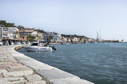Zakinthos, Greece - 01 August 2026: View of the waterfront in Zakinthos with boats moored along the stone promenade, buildings lining the coast, and clear blue water.