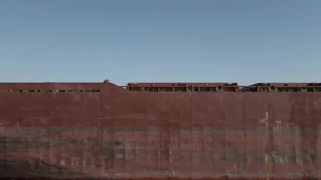 Rusting hull of a large cargo ship docked at port, under a clear blue sky.