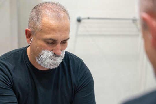 Handsome white-haired beared man shaving off his beard looking in a mirror. Concept of changing style