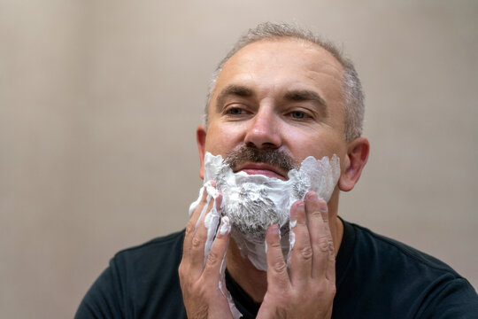 Portrait of handsome white-haired beared middle-aged man applying shaving foam to trim his beard