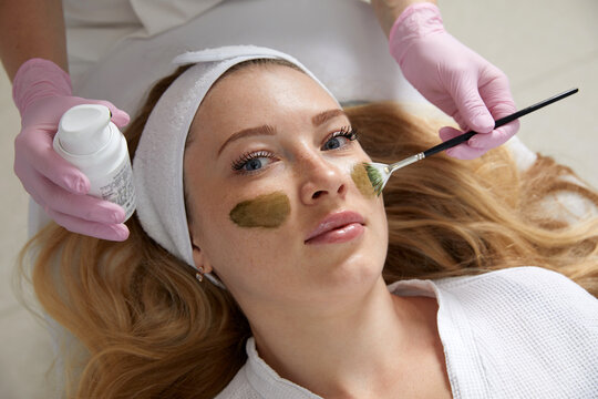Cosmetologist applying a mask from clay to the face of a beautiful woman. Spa treatment and face care in the beauty salon