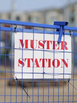 Muster Station safety sign attached with zip ties to blue wire fence