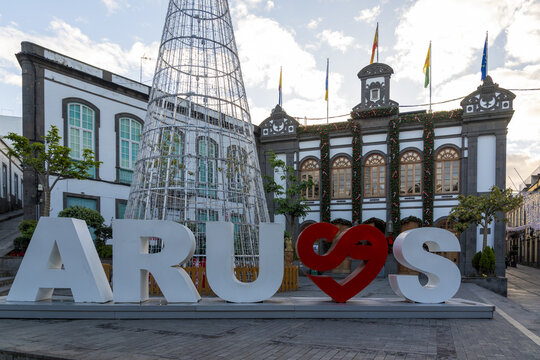 Arucas, Spain - 27 December 2025: View of the Arucas town sign with a heart symbol and an illuminated Christmas tree in front of the historic town hall decorated for the holidays.