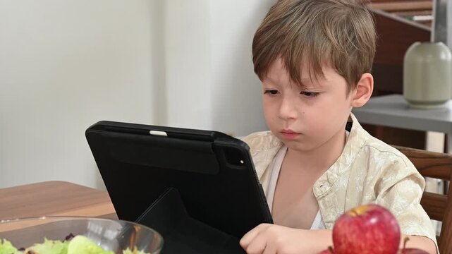 Young boy focused on tablet at kitchen dining table with salad bowl and apple nearby natural indoor light casual clothing calm concentration child using digital device