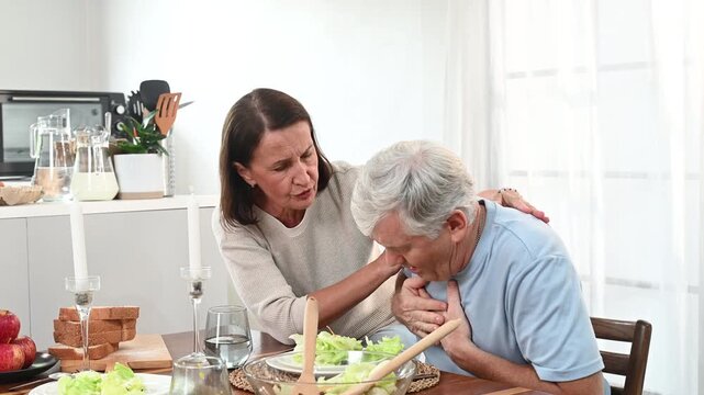Elderly man clutching chest with apparent heart attack pain as concerned woman comforts him at dining table offering water and support in emergency domestic scene of distress and concern