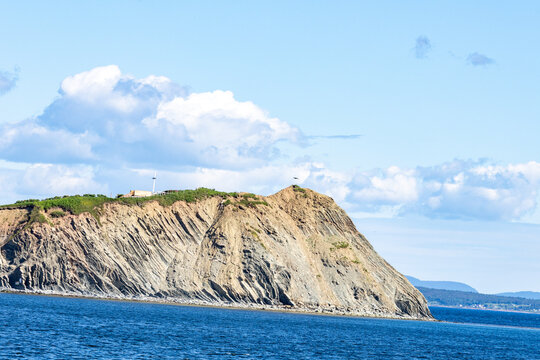 Gaspesie, Quebec, 15 July 2026 : A large rocky cliff rises from the deep blue ocean under a bright sky with scattered clouds.