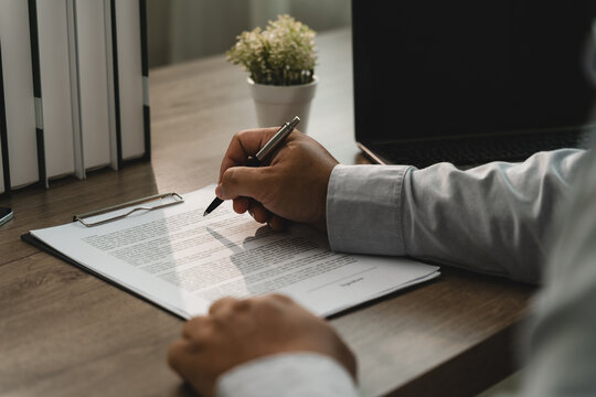 Business person checking a printed document with a pen on a clipboard beside a laptop, showing paperwork verification, office record review and a formal administrative checking process.