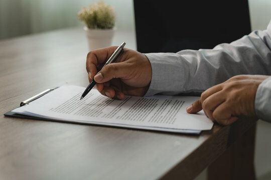 Close-up of hands using a pen to review printed paperwork on a clipboard at a desk, showing document verification, contract terms review and an office approval workflow with copy space.