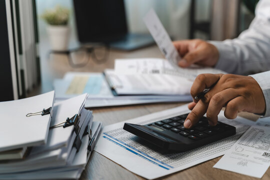 Hands using a calculator while holding printed financial paperwork at a busy office desk, showing expense review, cost calculation and a detailed financial document checking workflow.