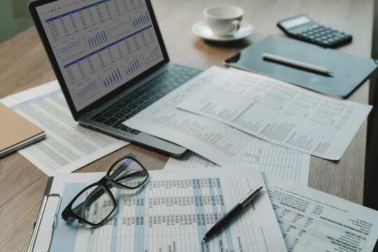 Laptop displaying financial dashboard beside printed spreadsheets, eyeglasses, pen, calculator on office desk, supporting document review, financial data analysis, reporting workflow.