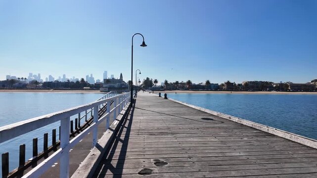 Wooden boardwalk of Kerferd Road Pier toward the coastline, revealing the distant Melbourne city skyline, beach foreshore, vintage light posts and clear sky in Albert Park, Victoria, Australia.