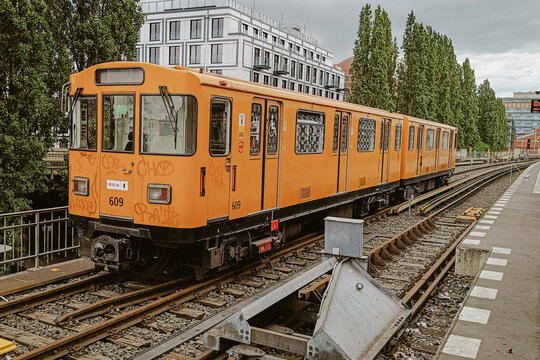 Berlin, Berlin, Germany - 05 June 2026: View of an orange Berlin U-Bahn train parked at a station, featuring graffiti on its side, with modern buildings and trees in the background.