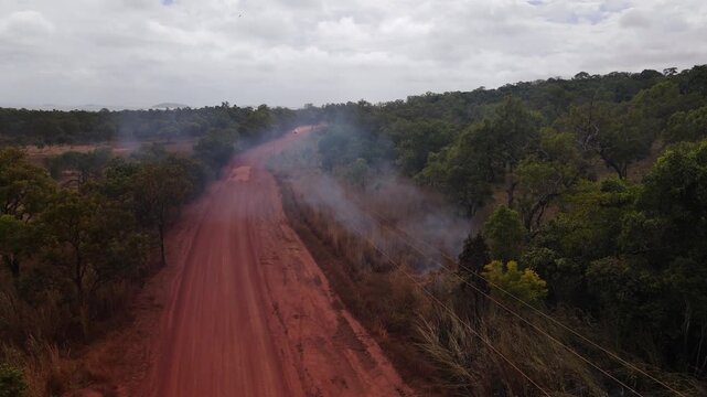 Static view of vehicle travelling along a dusty red dirt road in remote outback Australia, with a small grass fire burning beside the road. Regional transport, bushfire risk, remote landscape concept