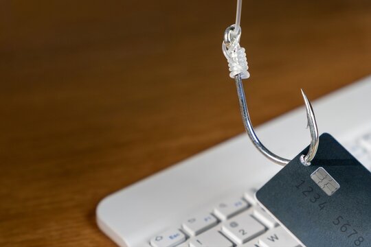 Fishing hook piercing a credit card over a blurred laptop keyboard. Online phishing scams, cybersecurity threats, digital fraud, and internet identity theft concept.