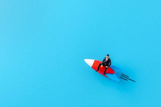 Miniature businessman figurine riding a rocket ship across a blue background. Copy space for text. Business launch, career advancement, startup success, financial growth concept