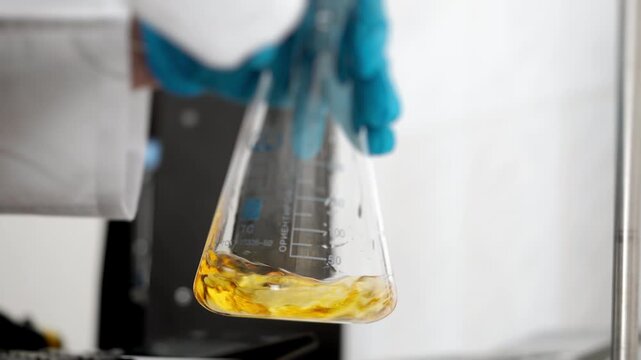 Laboratory worker swirls an Erlenmeyer flask in circular motion to mix yellow liquid during scientific research, chemical analysis and testing.