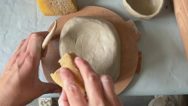 A woman shapes a handbuilt clay bowl on a round wooden board, smoothing the surface with a sponge and wooden tool, a handmade pottery craft process.