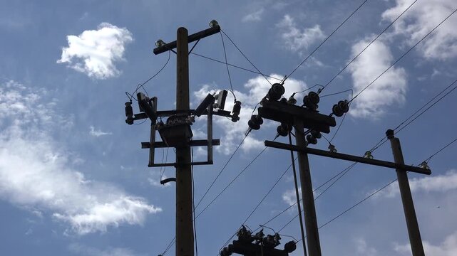 Concrete utility poles with a pole-mounted transformer, fuse cutouts and ceramic insulators seen from below against a bright cloudy sky. Static low-angle shot of an overhead power line