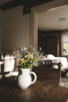 Laane County, Estonia - 25 May 2023: View of a white ceramic pitcher filled with blue and yellow wildflowers on a rustic wooden table in a cozy interior.