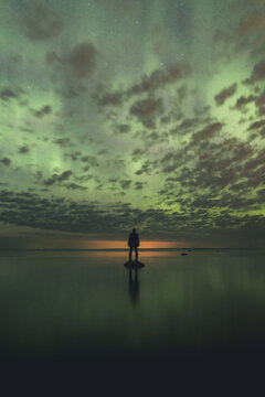 Merikula, Estonia - 23 April 2023: View of a person standing on a rock in the calm water under a vibrant display of the aurora borealis in the night sky.