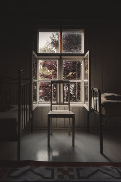 Laane County, Estonia - 08 June 2023: View of a wooden chair placed in front of an open window in a rustic room with metal bed frames and wooden walls, looking out towards lush trees.
