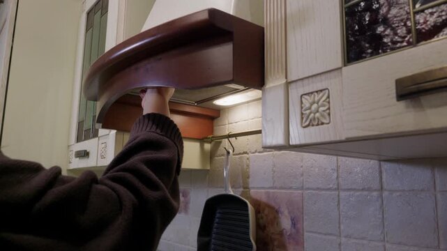 Person in black sweater reaches up to deactivate brown wooden range hood above stovetop in residential kitchen with tiled backsplash.