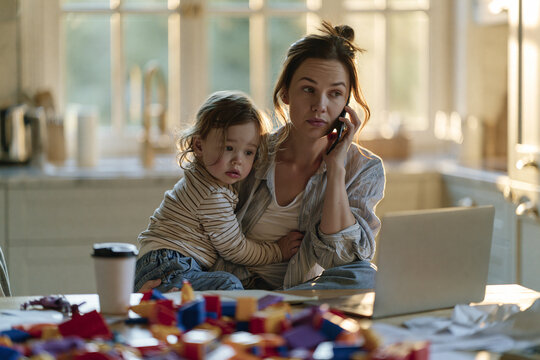 A busy mother juggling work and childcare, talking on the phone with her child on her lap. A laptop and toys are scattered on the table, highlighting the chaotic yet loving atmosphere