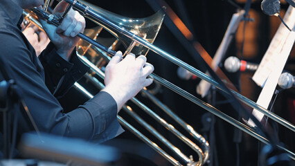 Closeup of hands gripping trombone slide under stage lighting. Reflective brass surface, dark clothing, blurred background with microphones © VeNN