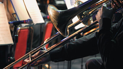 Closeup of trombone player during symphony orchestra performance. Warm stage lighting highlights brass instrument hands. Music stands with sheet music visible in background © VeNN