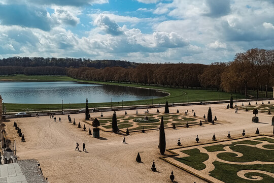 Paris, France - 02 April 2026: View of the formal gardens at the Palace of Versailles with manicured hedges, gravel paths, and a large pond under a cloudy sky.