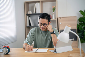 Obraz premium Thoughtful man wearing glasses sitting at wooden desk writing on blank paper with tablet stand and modern lamp in bright home office space