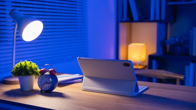 Modern workspace with digital tablet, alarm clock, desk lamp, potted plant, notebook, and cozy ambient lighting for nighttime productivity