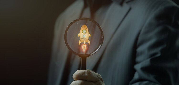 Close-up shot of hand holding magnifying glass to inspect glowing rocket launch target icon inside display. Startup business growth acceleration and project launch strategy inspection concept.