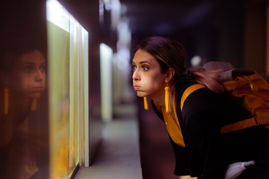 Curious Young Woman Traveler Looking Closely Inside Underwater Zoo Aquarium. Interested female tourist with backpack examines glowing fish tank reflecting her unique face