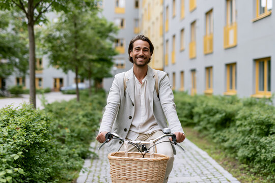 Man smiles while cycling through green urban path. Basket adds charm to his casual ride. Modern building lines softly frame the background. His joy suggests a relaxed, everyday commute