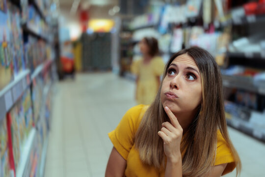 Thoughtful Young Woman Looking Up Confused While Shopping In Supermarket. Puzzled female customer stands inside grocery store aisle choosing products with hand on chin 