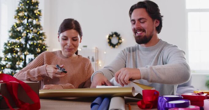 Christmas couple wrapping surprise gift with ribbon and gold paper at home near festive Xmas tree. Happy woman with help of cheerful man unrolling wrapper and cutting paper with scissors to wrap box