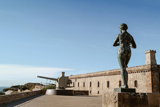Barcelona, Spain - 05 December 2025: View of the historic Montjuic Castle featuring a bronze statue in the foreground and a large coastal defense cannon against a clear blue sky.