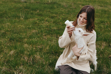 Obraz premium A woman in a cozy knitted sweater feeds a white baby goat with a bottle in a countryside field, capturing a warm lifestyle moment and gentle animal bonding.