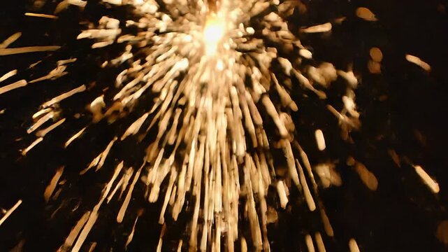 Golden fireworks burst expands across the night sky, showing a bright celebratory explosion developing from initial flare to scattering sparks.