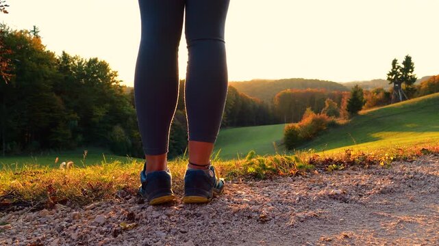 CLOSE UP, LENS FLARE: Back view of a woman standing on scenic hill at golden hour, with the sun shining between her legs. She stopped while running to admire autumn forest, rolling hills and meadows.