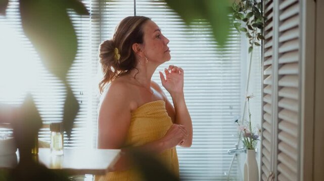 Side view of a mature woman smiling at her reflection and touching her face in the bathroom, showing confidence, self care and satisfaction with her appearance.