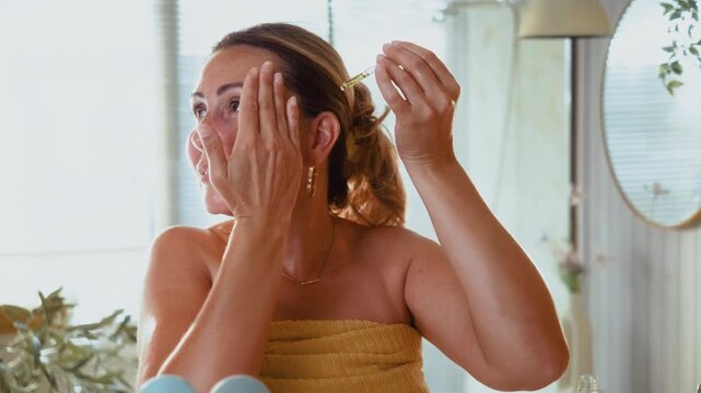 Smiling mature woman applies facial oil with a pipette in the bathroom, enjoying a healthy skincare routine and confident self care at home.